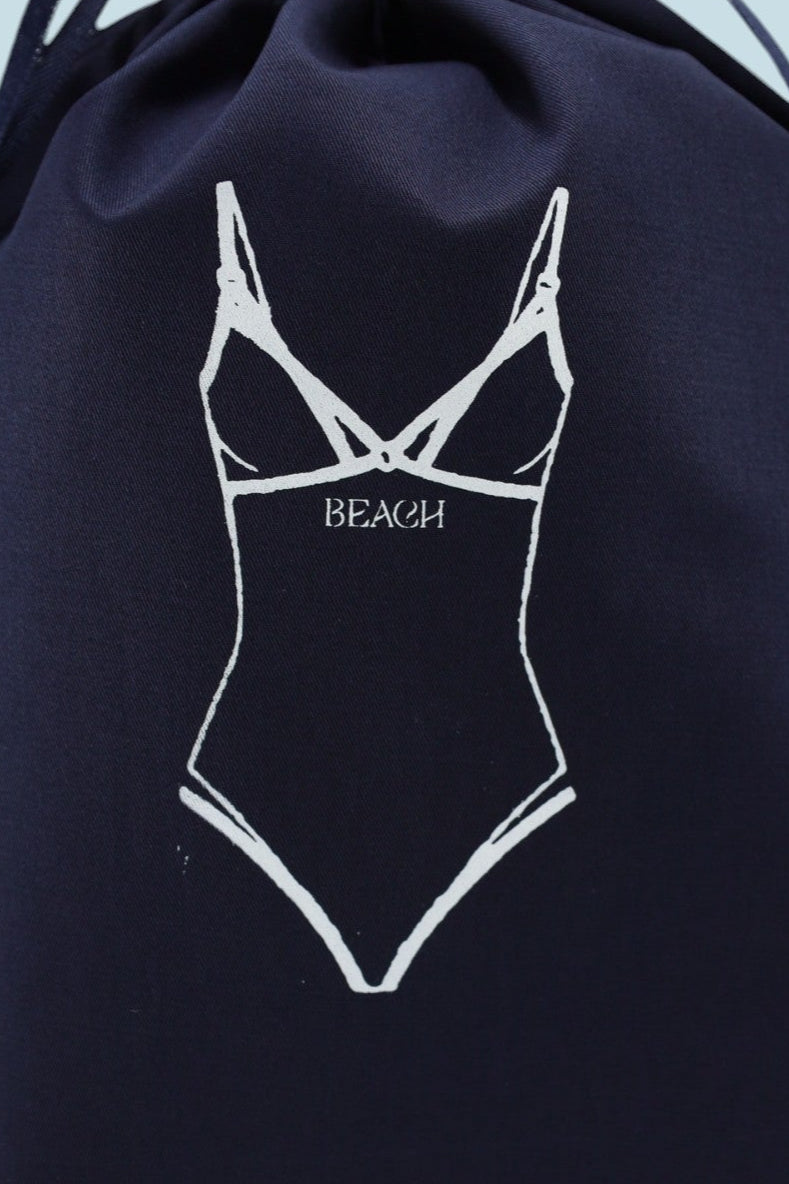 Pochon Moonlac en coton bleu marine avec sérigraphie blanche d’un maillot de bain une pièce féminin, inscription “BEACH”, posé debout