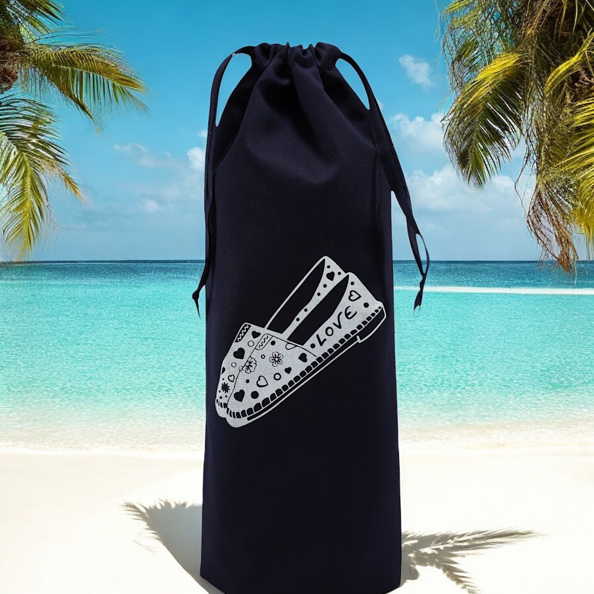 Pochon bleu marine Moonlac avec dessin d’espadrilles, posé sur le sable avec la mer en arrière-plan – ambiance estivale et vacances assurées.