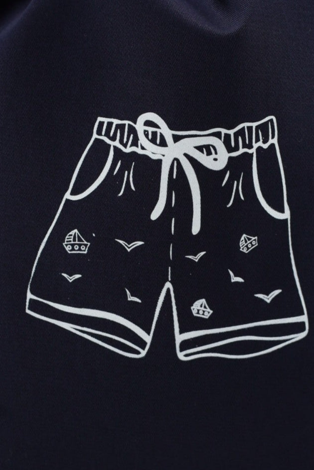 Pochon Moonlac en coton marine, sérigraphié d’un short de bain blanc, posé debout 